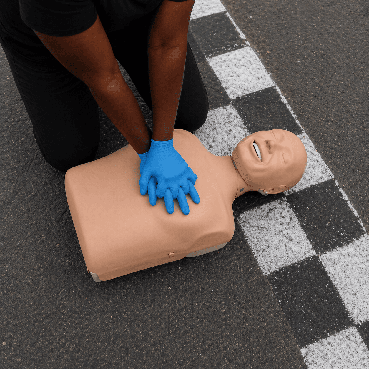 A CPR training dummy lying on a checkered race track line while a woman wearing blue gloves performs chest compressions.